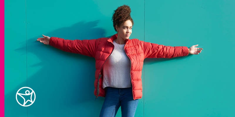Young smiling female student in a red coat wit her arms against a turquoise wall.