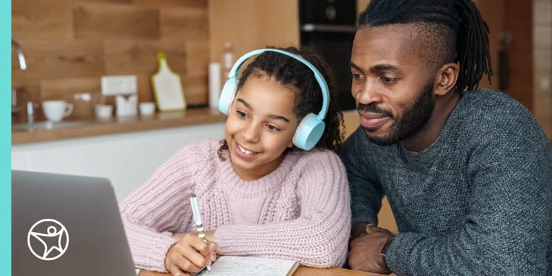 A young girl in a pink sweater and turquoise headphones is sitting at her laptop with her father in a grey sweater working togther on her Connections Academy online class.