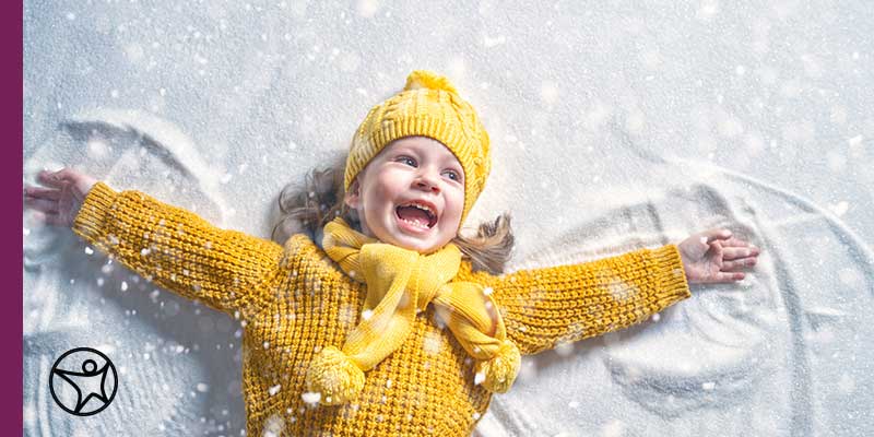 An elementary student wearing a bright yellow hat and sweater making an angel in the snow.