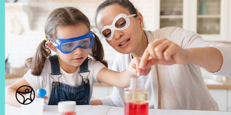 A young female student in denim overalls working with her mom/learning partner on a science experiment.