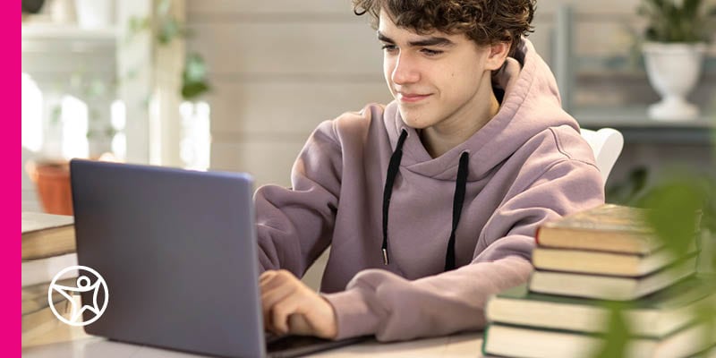 A teenage student sitting at a desk and looking at their laptop which is open next to pile of books
