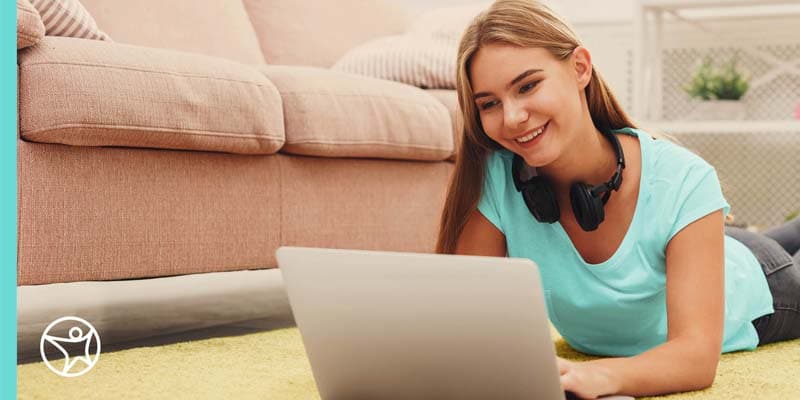 A high school student wearing a bright blue shirt sitting on the floor working on her laptop
