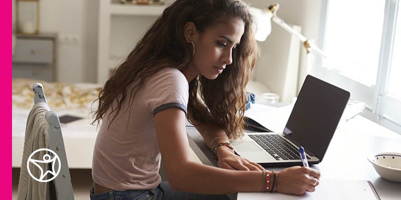An online student in a white t-shirt working by her laptop taking notes for a Connections Academy class.