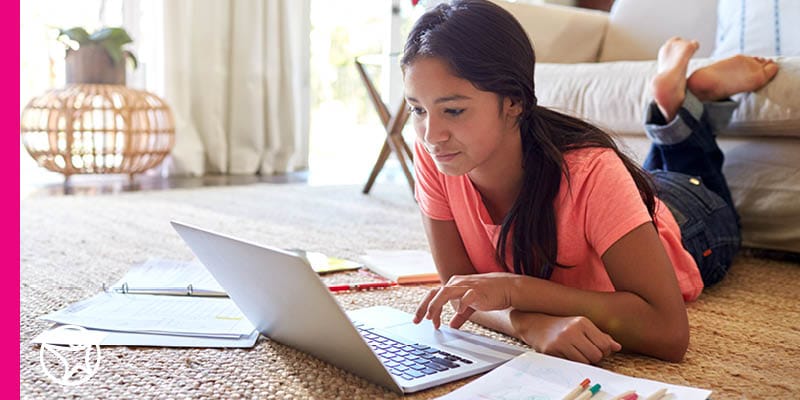 Young teen in an orange shirt is laying on the floor in her living room working on a laptop for an online class at Connections Academy for a career program during the afternoon.