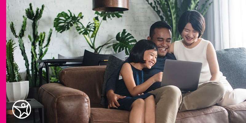 Three individuals sitting on a brown leather couch, looking at a laptop placed on one person's lap. They are in a living room with a modern decor, including a white brick wall, lush green plants, and a sleek black desk with a computer in the background. The room is well-lit with natural light complemented by a hanging lamp above.