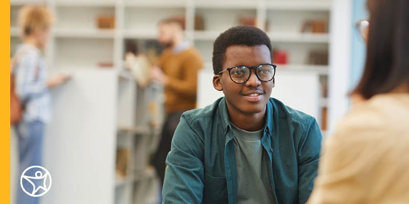 Young male teen tudent in a denim shirt talking to a an adult in a yellow shirt with glasses.