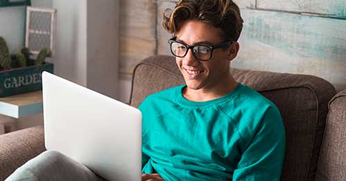 Young student in a green shirt working on an online school class at home sitting on a brown sofa.