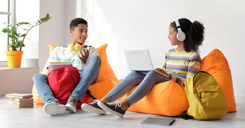 2 young students working together during their online classes sitting in orange bean bags. 