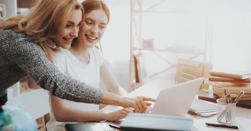 Image of a young student in a white shirt looking at a laptop while taking some pointers from her mom. 