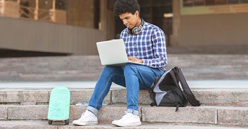 An online student sitting on steps outside while researching college websites for high school students on a laptop.