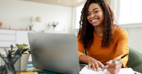 Image of a young female Connections Academy student in ayellow shirt sitting down smiling looking at their laptop. 