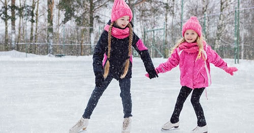 Two young females on an ice rink in pink coats ice skating and enjoying a snowy winter day. 