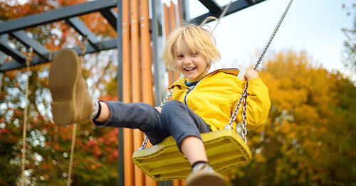 Image of a young female Connections Academy student in a yellow coat swinging at the park during a fall day. 