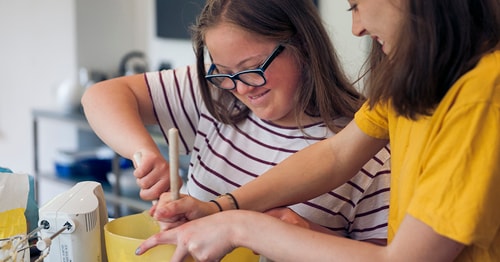 Two teens working together in a kitchen baking a cake. One teen is wearing a yellow shirt, and the other teen is wearing a striped shirt. 