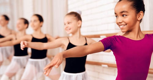 A group of young female ballet students practicing at the barre. One is wearing pink and the rest are wearing a black leotard.