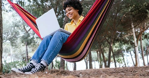 An online student sitting on a red striped hammock working on their online class outside under a tree. 