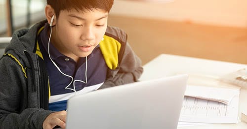 A student wearing a grey hoodie and a blue and yellow shirt with headphones sitting at a laptop working on an online assignment for Connections Academy. 