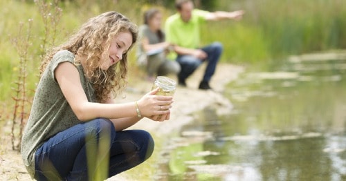 Young student in a green shirt is looking at pond water in a mason jar. 