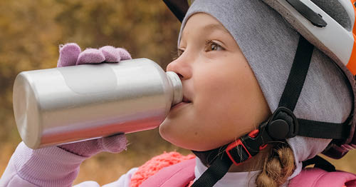 Image of a young female Connections Academy student in a pink dress wearing a bicycle helmut and is drinking a metal container full of water.  