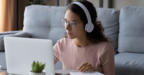 Image of a young female Connections Academy student in a pink sweater wearing white headphones looking at her laptop. 