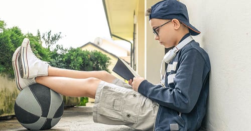 Image of a young male Connections Academy student sitting against a wall reading a book. 